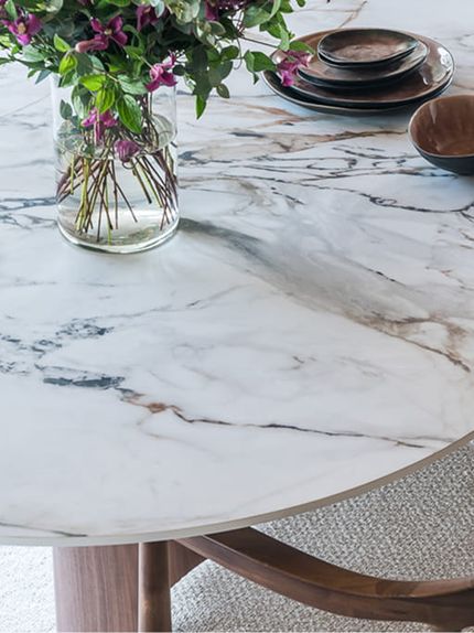 Close-up van een afgeronde eettafel met een wit marmerlook tafelblad en uitgesproken grijze en bruine aders, gedecoreerd met een vaas bloemen en servies.