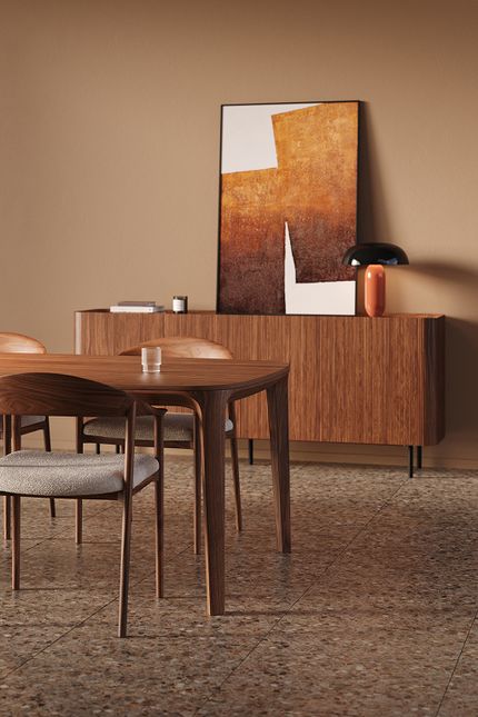 Close-up van een warm houten dressoir met verticale latjes, met grote abstracte poster en tafellamp; op de voorgrond een houten eettafel met stoelen.