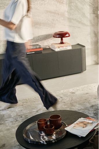Une personne passe devant un buffet sombre contre un mur en béton, avec une pile de livres et une lampe de table bordeaux ; au premier plan, une table basse ronde.