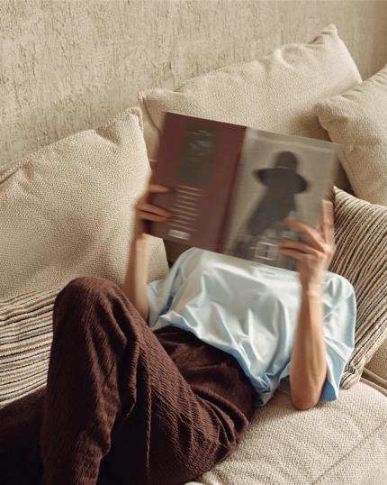 Persoon ligt op een lichtbeige zetel met grote kussens en leest een boek; gestreept kussen en warme muur op de achtergrond.