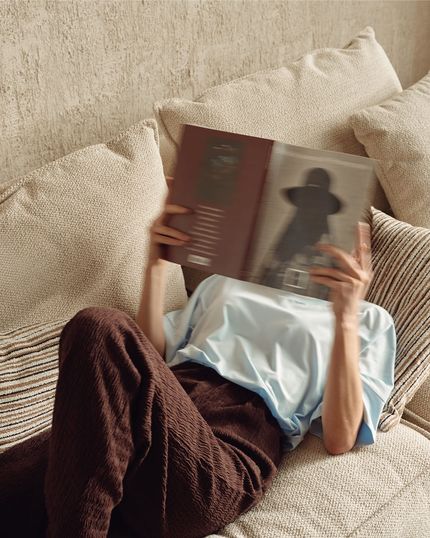 Persoon ligt op een lichtbeige zetel met grote kussens en leest een boek; gestreept kussen en warme muur op de achtergrond.