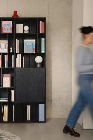 Boekenkast met boeken en objecten in een minimalistisch interieur, met een persoon die door de ruimte beweegt.