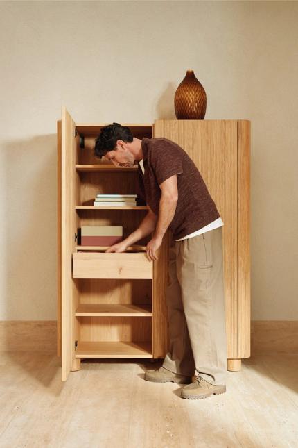 Man opent een hoge houten kast met verticale latjes en schuiflade, met boeken op de planken en een vaas bovenop.