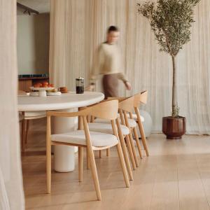 Une femme portant un pull beige marchant autour d'une table ronde avec des chaises en bois. Sur la table, il y a un bol de fruits et une tasse de café, et un olivier se trouve à droite.