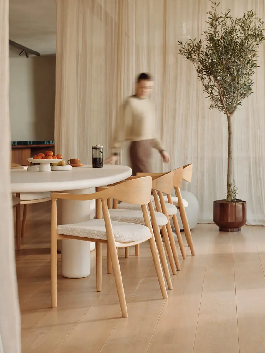 Moderne eetkamer met houten stoelen, ronde tafel en een olijfboom in een lichte, minimalistische ruimte.