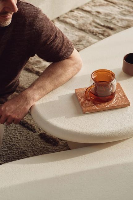 Close-up van een witte, organisch gevormde salontafel met amberkleurige karaf op onderzetter en een mok; persoon zit ernaast op een vloerkleed.