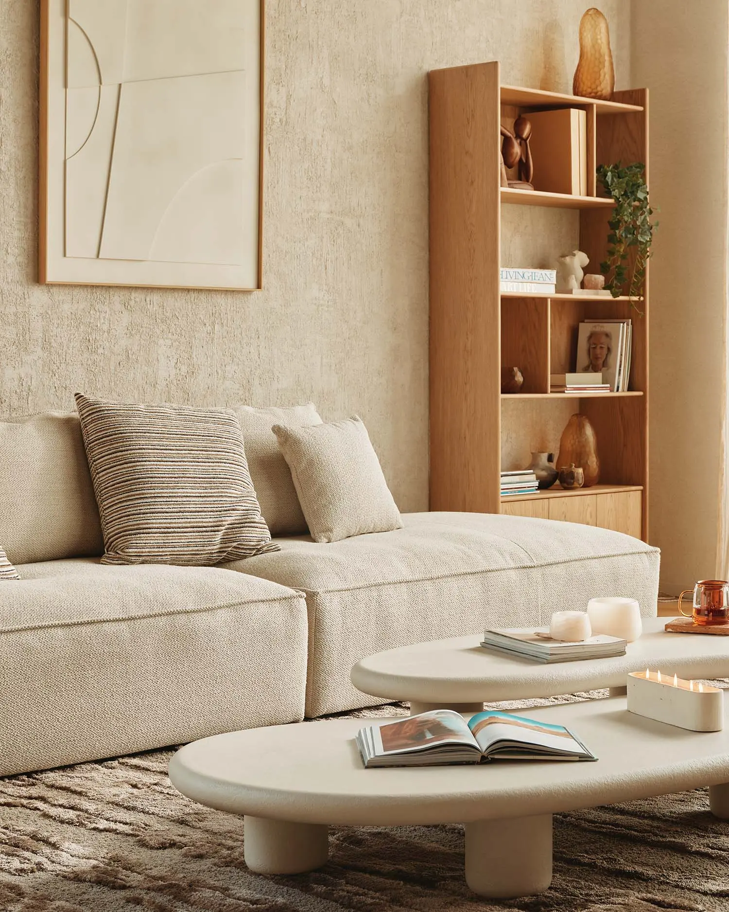 Lichte woonkamer met beige modulaire zetel, organische salontafels en houten boekenkast bij groot raam.