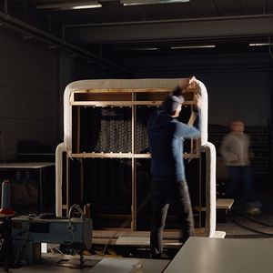 Vakman in een werkplaats die de bekleding van een bank vastmaakt. De bank heeft een houten frame en is gedeeltelijk bekleed met stof.