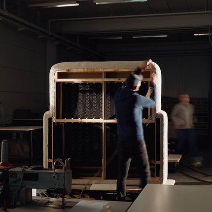 Vakman in een werkplaats die de bekleding van een bank vastmaakt. De bank heeft een houten frame en is gedeeltelijk bekleed met stof.