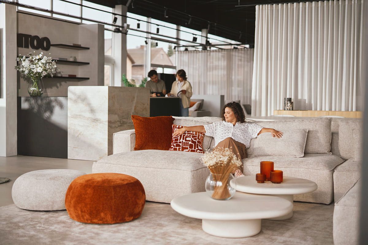 Showroom avec des canapés clairs, des coussins orange-rouge et des objets décoratifs, avec une personne se déplaçant dans l'espace.