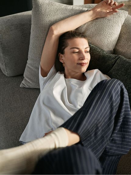 Une femme se détend sur un canapé en tissu gris avec des coussins, le bras au-dessus de la tête, baignée d’une lumière chaude.