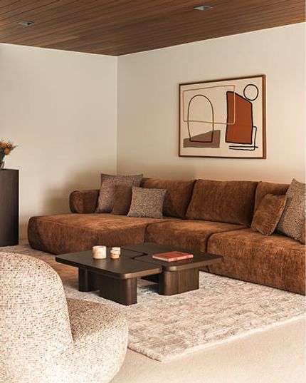 Warme woonkamer met een bruine hoekzetel, donkere salontafel en fauteuil in neutrale tinten.