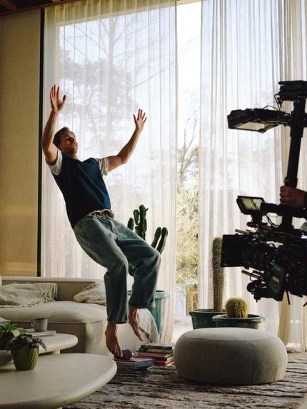 Coulisses dans un salon : un homme saute près d’un canapé clair tandis qu’un cadreur filme la scène avec un rig devant de grandes fenêtres.