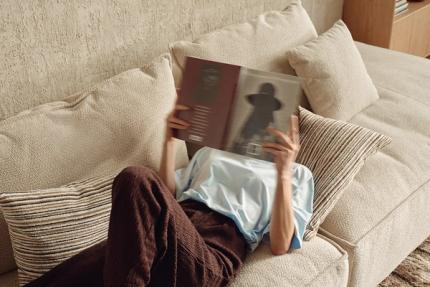 Personne allongée sur un canapé en tissu beige, lisant un livre, avec des coussins dans des tons neutres et doux.