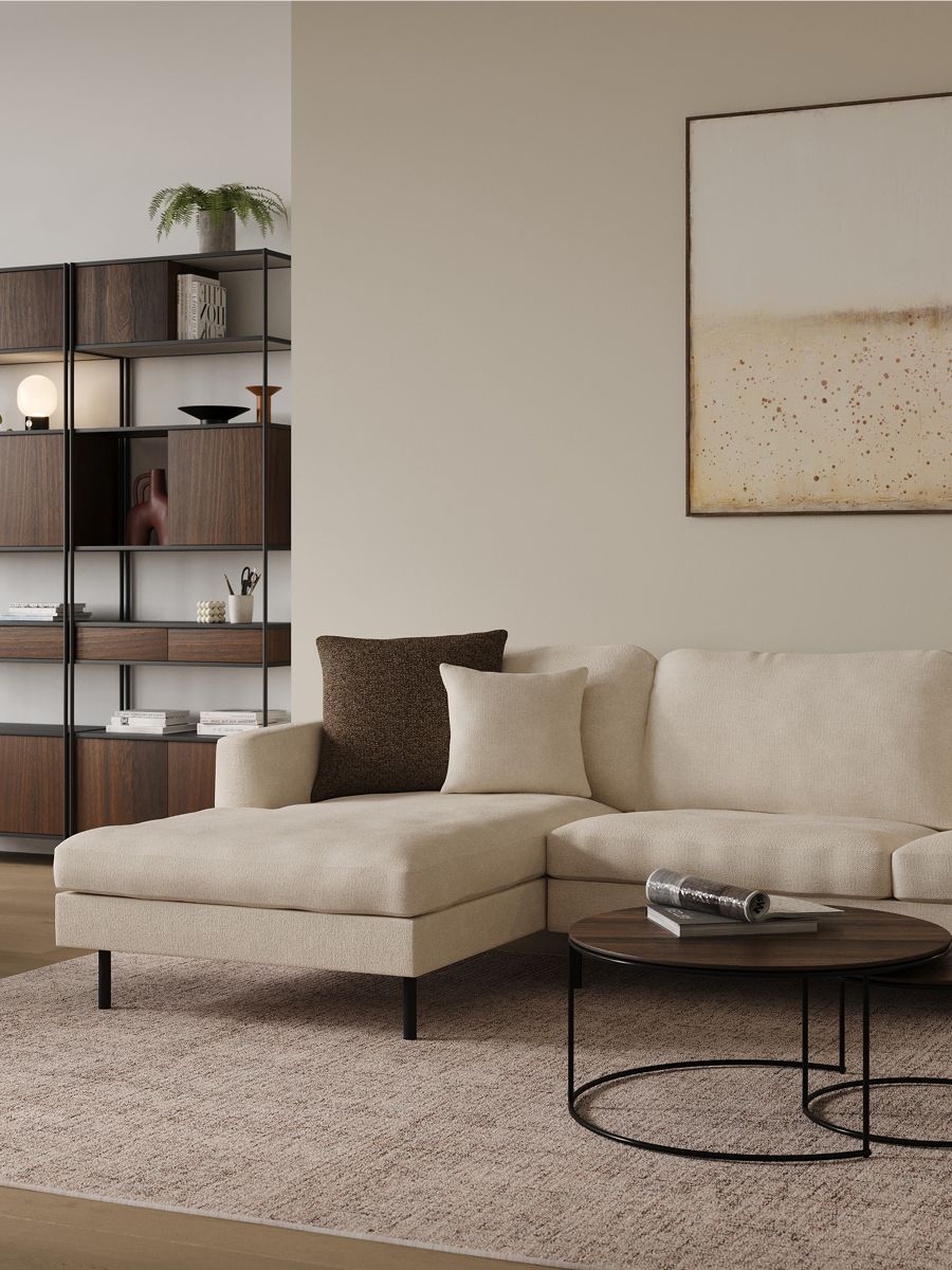 Detail van een beige hoekzetel met kussens, ronde zwarte salontafel en open kast met donkere houtvakken en planten.