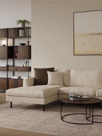 Détail d’un canapé d’angle beige avec coussins, table basse ronde noire et étagère ouverte en bois foncé avec plantes.