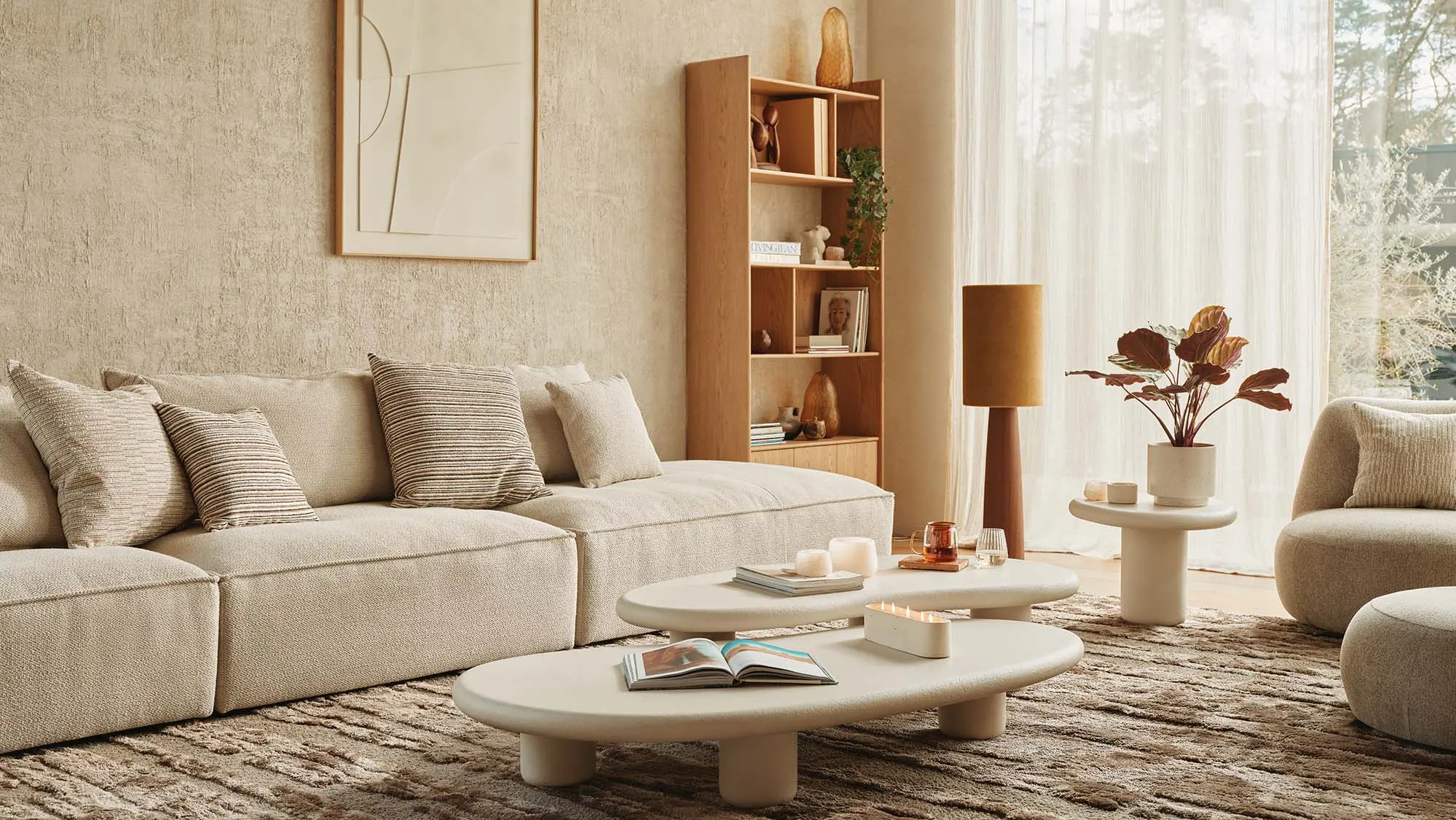 Lichte woonkamer met beige modulaire zetel, organische salontafels en houten boekenkast bij groot raam.