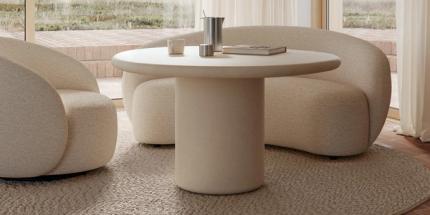 Lichte zithoek met ronde tafel, twee zachte zetels en hanglampen, naast een hoge houten kast en grote ramen met gordijnen.