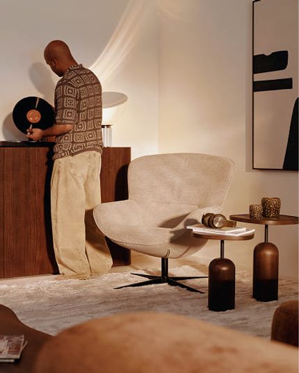 Persoon kiest een vinylplaat naast een lichte fauteuil en bijzettafels in een warm interieur.