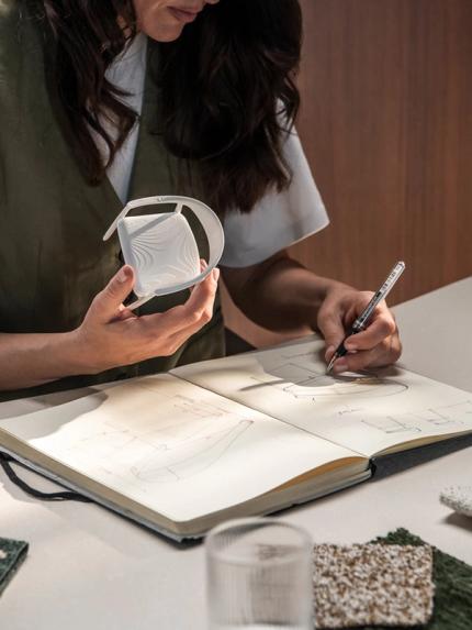 Persoon die een 3D-geprint object vasthoudt en ontwerptekeningen maakt in een notitieboek, met stoffen en glazen voorwerpen op de tafel.