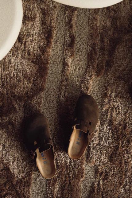 Bruine leren slippers op een zacht, beige tapijt, met een poef zichtbaar aan de rechterkant.