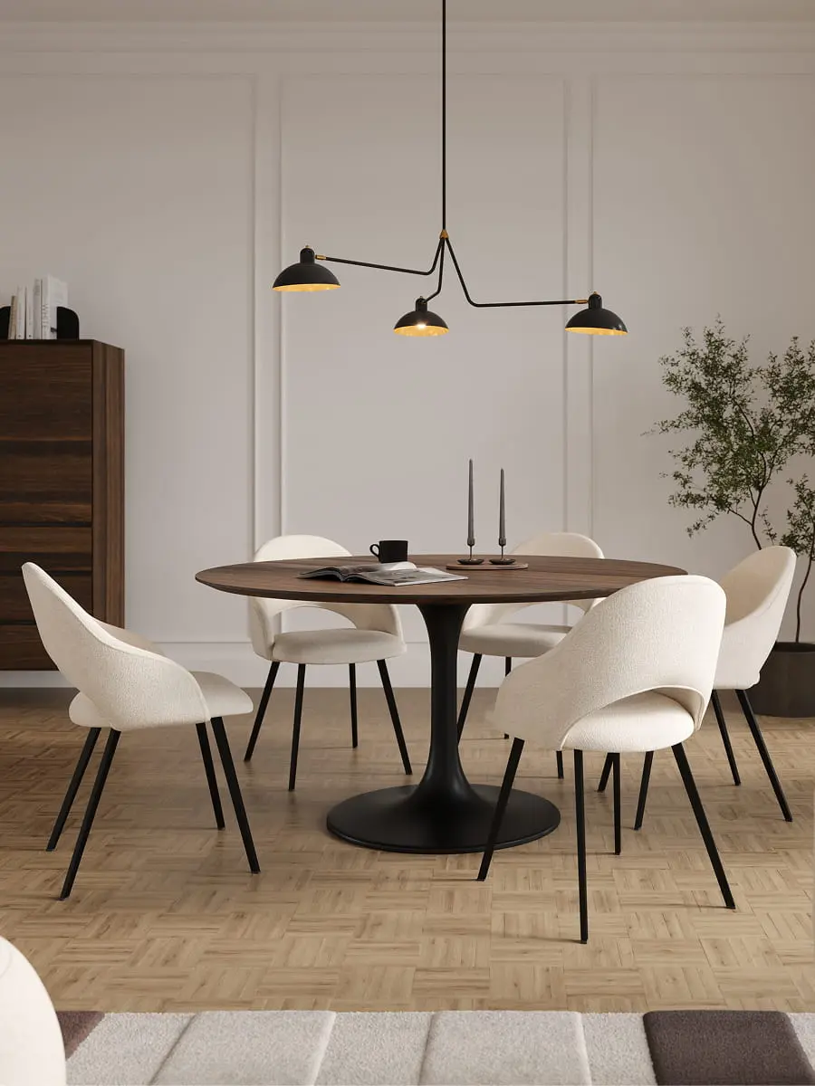 Eetkamer met ronde houten tafel op zwart metalen voet, omringd door lichte stoelen met zwart metalen poten. Op tafel liggen kaarsen, een kopje en een open boek, met een groene plant op de achtergrond.