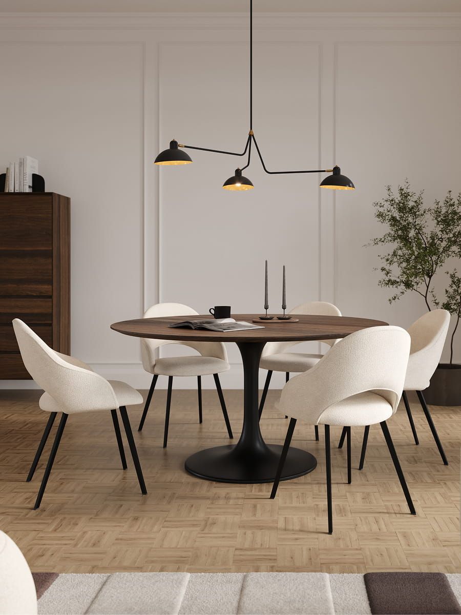 Eetkamer met ronde houten tafel op zwart metalen voet, omringd door lichte stoelen met zwart metalen poten. Op tafel liggen kaarsen, een kopje en een open boek, met een groene plant op de achtergrond.