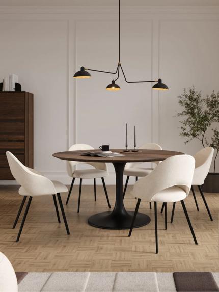 Eetkamer met ronde houten tafel op zwart metalen voet, omringd door lichte stoelen met zwart metalen poten. Op tafel liggen kaarsen, een kopje en een open boek, met een groene plant op de achtergrond.