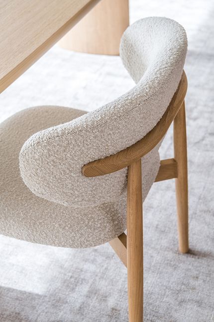 Close-up van een eetkamerstoel met zachte, lichtbeige bouclé bekleding en afgeronde armleuningen op een houten frame, naast een houten tafel.