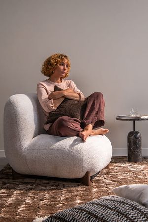 Persoon zit ontspannen met gekruiste benen in een afgeronde, licht gestoffeerde fauteuil en houdt een donker kussen vast, naast een marmeren bijzettafel.