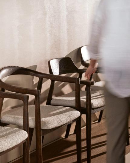 Moderne houten eetkamerstoelen met beige kussens, tegen een lichte muur, met een persoon die door de ruimte beweegt
