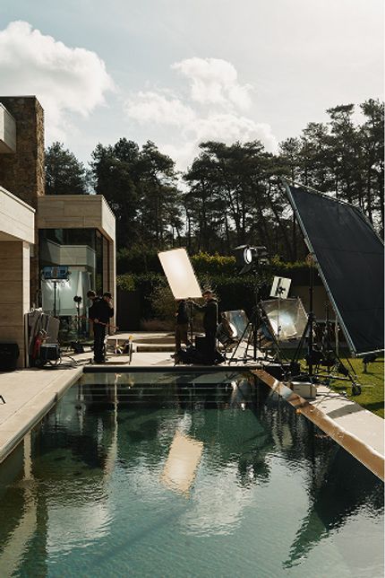 Behind-the-scenes filmset bij modern huis met zwembad; crew en grote reflectieschermen staan opgesteld in de tuin.