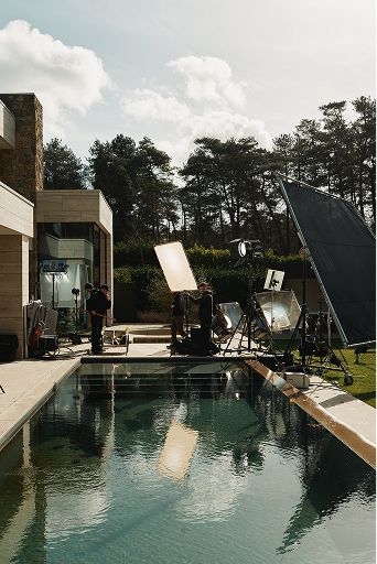 Behind-the-scenes filmset bij modern huis met zwembad; crew en grote reflectieschermen staan opgesteld in de tuin.