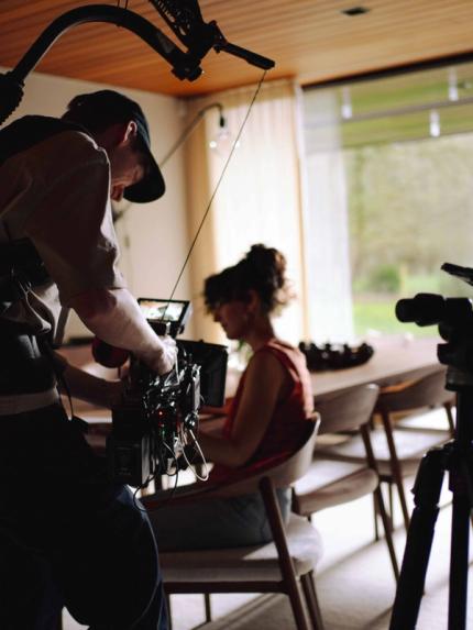 Un caméraman filme avec un rig en intérieur; à l’arrière-plan, une personne assise près de la fenêtre, floue.