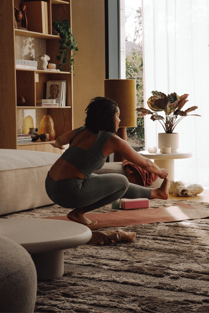 Vrouw stretcht op yogamat in woonkamer terwijl cameracrew met filmcamera en lampen de scène opneemt.