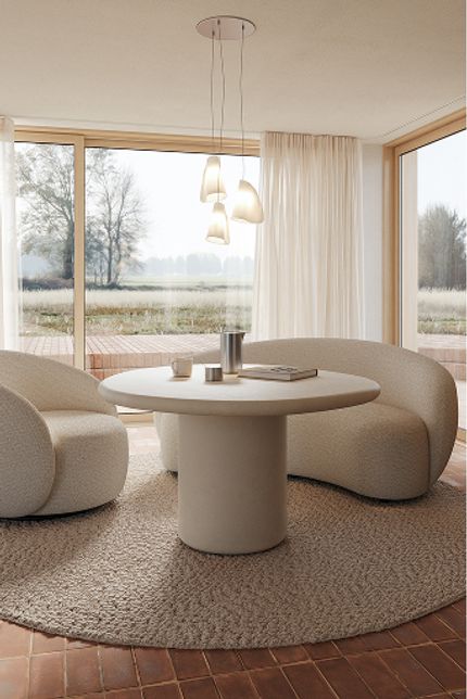 Ronde bijzettafel met zachte, organische fauteuils op een rond tapijt in een lichte zithoek met grote ramen en transparante gordijnen.