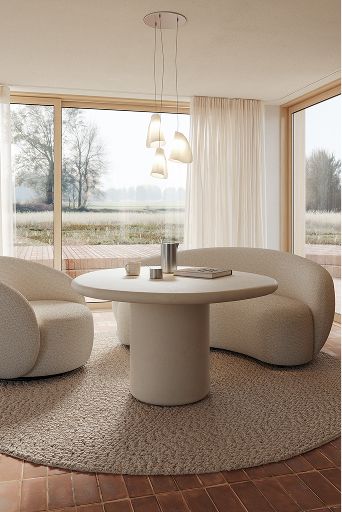 Ronde bijzettafel met zachte, organische fauteuils op een rond tapijt in een lichte zithoek met grote ramen en transparante gordijnen.