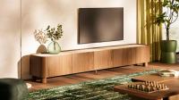 Meuble TV long en bois avec coins arrondis sous une télévision murale, avec un vase vert et la lumière chaude du soleil entrant.