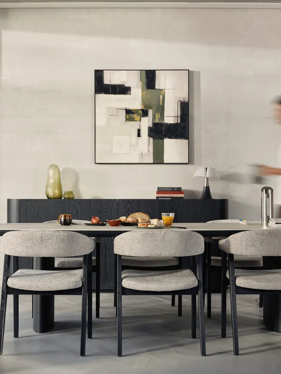 Lange eettafel met vier grijze stoelen en donkere poten, stilleven op tafel, abstract kunstwerk aan de muur en persoon in beweging.