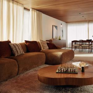 Canapé Sarpi en tissu brun chaleureux dans un salon lumineux, avec une table basse ronde en bois et un échiquier, et un coin repas à l’arrière-plan.