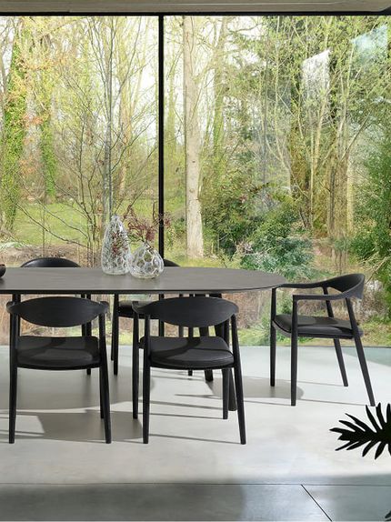 Moderne eetkamer met een lange, donkere eettafel en zwarte houten stoelen, voor grote ramen met uitzicht op een groene, bosrijke tuin.