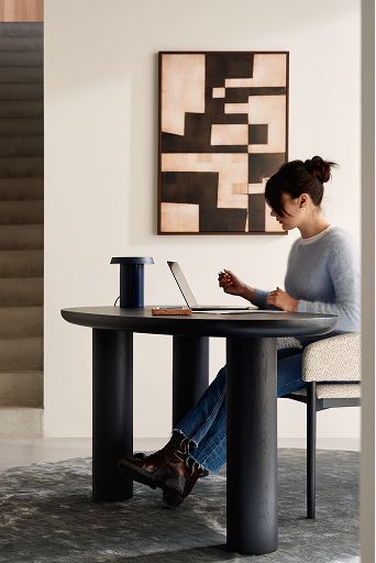 Persoon werkt aan zwarte ovale consoletafel met laptop; trap en abstracte kunst aan de muur.