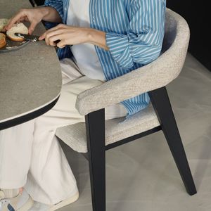 Detail van Gubbio stoel: lichtgrijze bekleding met armleuning en zwart houten onderstel, met iemand aan tafel.