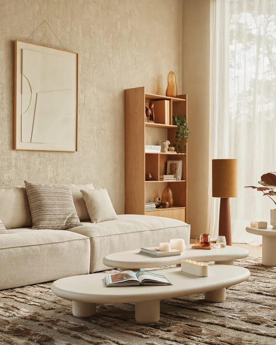 Een stijlvolle woonkamer met een beige bank, twee organisch gevormde salontafels met boeken en kaarsen, een houten boekenrek met decoratieve objecten en een abstract kunstwerk aan de muur, alles in een warme, minimalistische stijl.