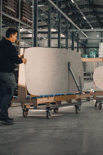 Medewerker in een productiehal verplaatst grote, lichtgekleurde tafelbladen met afgeronde en organische vormen op verrijdbare rekken.
