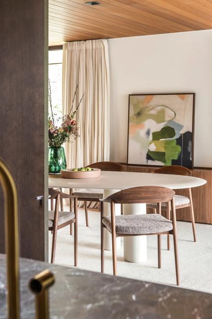 Witte ovale eettafel met houten stoelen, groene vaas met bloemen en abstract schilderij in lichte eetkamer.
