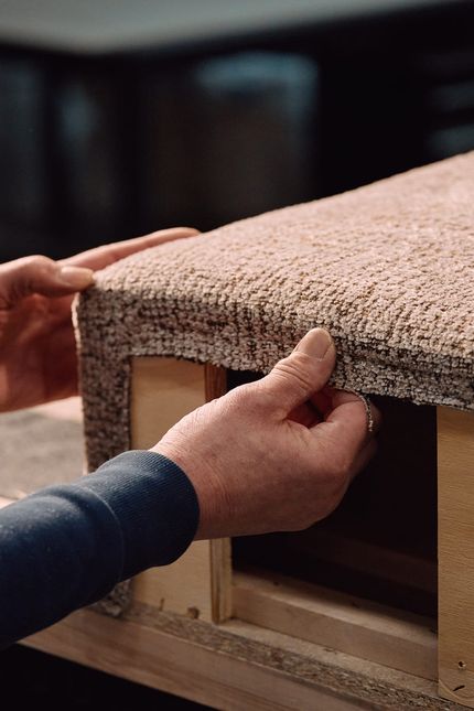 Close-up van handen die een bekleding in structuurstof strak trekken over het houten frame van een zitmeubel.