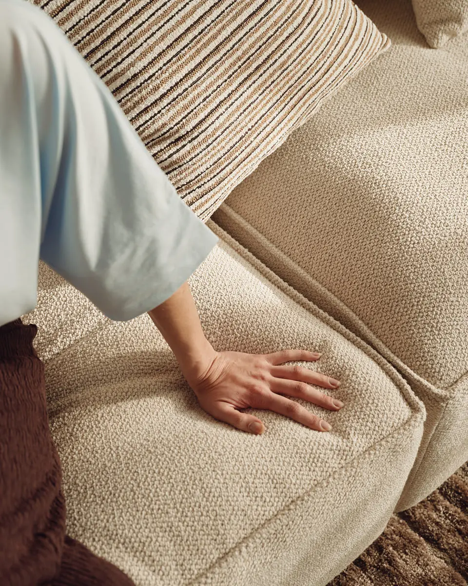 Close-up van een hand op de zitting van een lichtbeige zetel in structuurstof, met gestreept kussen in warm daglicht.