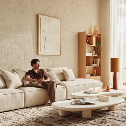 Moderne zithoek met een beige bank, een salontafel van licht materiaal en een boekenrek op de achtergrond. Een man zit ontspannen op de bank, genietend van het interieur.
