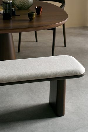 Gros plan d’un banc de salle à manger avec une assise rembourrée claire et un piètement en bois foncé, placé devant une table à manger arrondie en bois foncé.